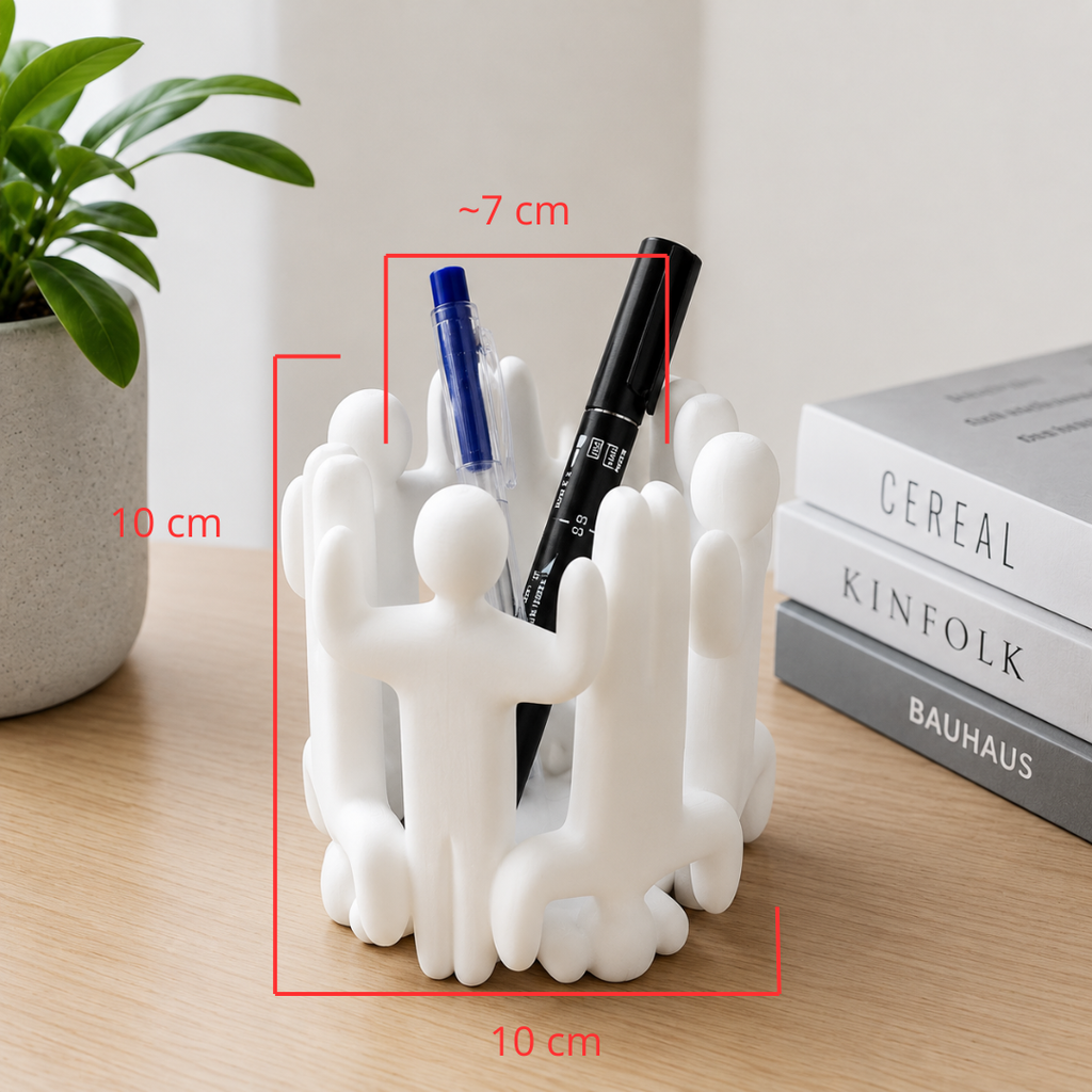 A white sculptural pen holder shaped like stylized human figures sits on a wooden desk, with red dimension lines showing measurements of approximately 7 cm width and 10 cm height/depth. A plant and stacked books are visible in the background.